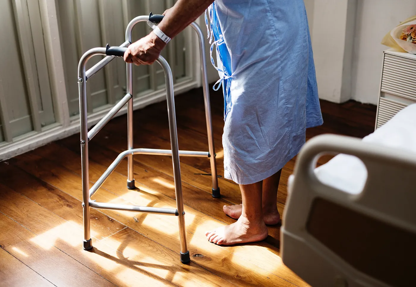 A person in a hospital gown standing with a walking frame beside a bed